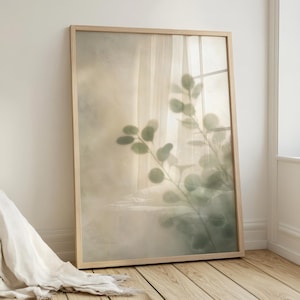 Può includere: Una stampa artistica incorniciata con un'immagine morbida e astratta di foglie di eucalipto in tonalità verdi e grigie. L'opera d'arte è racchiusa in una cornice di legno chiaro, creando un'estetica naturale e serena. La stampa è appoggiata a un muro.