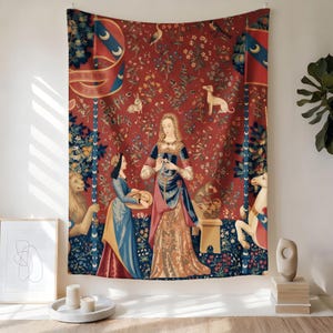 May include: A large tapestry with a red background featuring a medieval scene. The tapestry depicts women in long gowns, animals, and floral patterns. Two banners with crescent moon designs are on the top corners. The tapestry is hanging on a white wall.