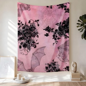May include: Pink tapestry with a gothic theme. Features black floral and bat illustrations, along with a spider web design. The bats are in flight, and the flowers are clustered. The tapestry is hanging on a wall.