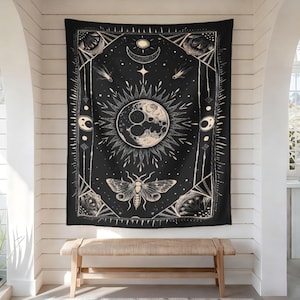 May include: Black tapestry with a celestial design featuring a sun, moon, stars, and a moth. The artwork is in cream and white, with a geometric border. The tapestry is hanging on a white wall above a wooden bench.