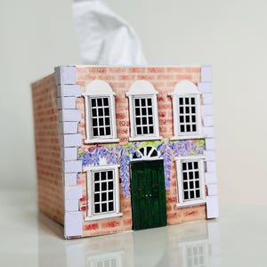 May include: A house-shaped tissue box cover with a brick facade, white-framed windows, and a green door. The design includes floral elements, and a white tissue is visible.