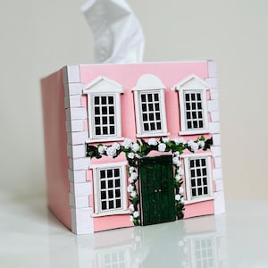 May include: A decorative pink tissue box designed as a house, with white window frames, a green door, and a garland of white roses. White tissues are visible. A charming home decor item.