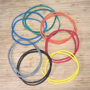 May include: A selection of colourful braided rope circles in shades of blue, green, orange, red, yellow, grey, and black. The ropes are arranged on a light brown wooden surface, creating a visually appealing pattern.