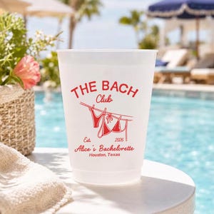 May include: A white plastic cup with red text that reads "THE BACH Club" and "Alice's Bachelorette Houston, Texas". The cup has a drawing of a bikini hanging on a clothesline. The background shows a pool and lounge chairs.
