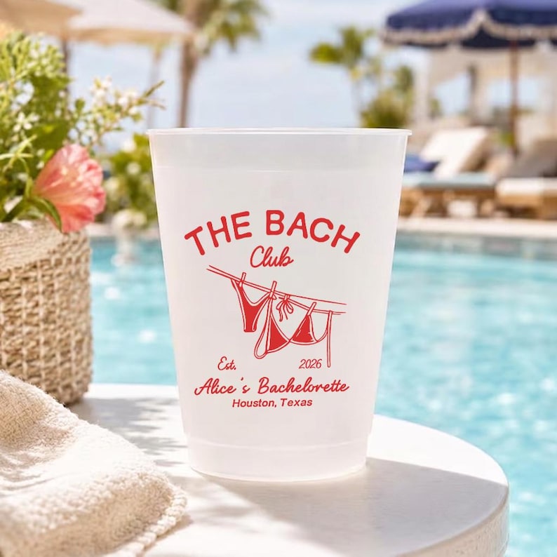 May include: A white plastic cup with red text that reads "THE BACH Club" and "Alice's Bachelorette Houston, Texas". The cup has a drawing of a bikini hanging on a clothesline. The background shows a pool and lounge chairs.