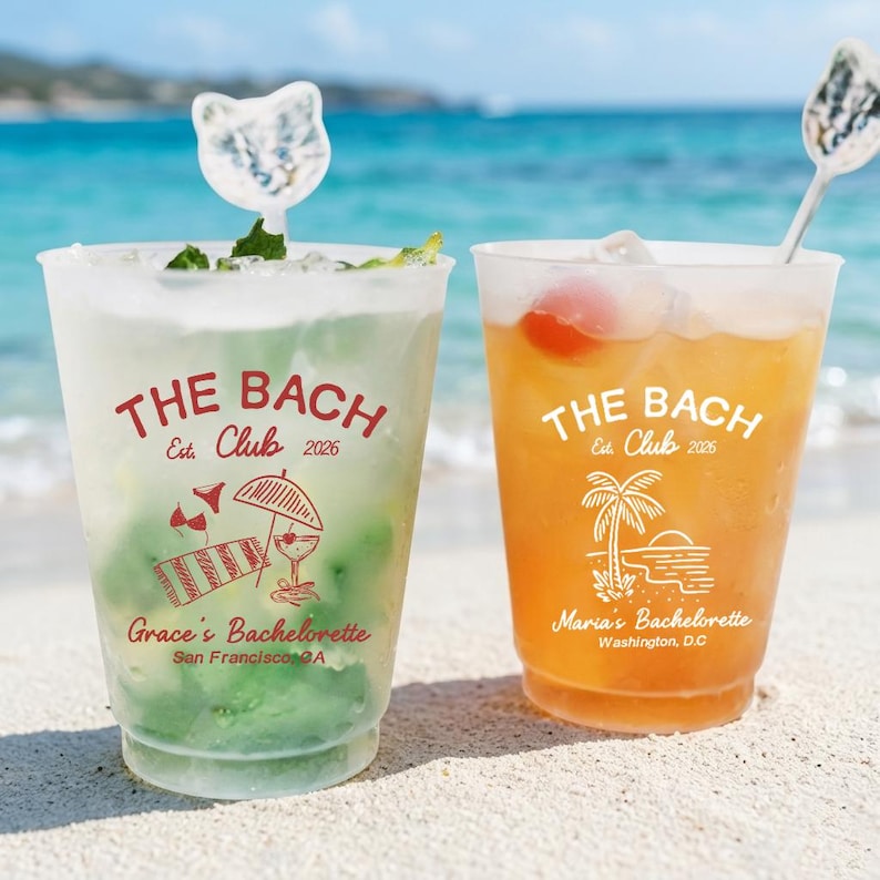 May include: Two clear plastic cups filled with cocktails, one green and one orange, on a sandy beach. The cups have the text "THE BACH Est. Club 2026" and personalized text. Each drink has a decorative stirrer.
