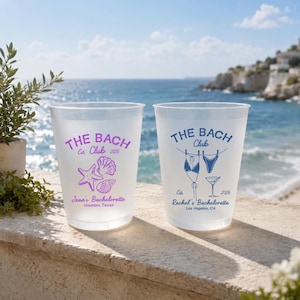 May include: Two frosted plastic cups with "THE BACH Club" printed on them. One cup has a purple seashell and starfish design, the other has a blue bikini and martini glass design. Both cups have the year 2026.