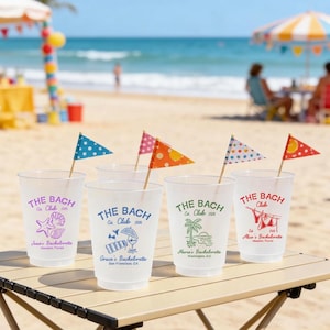 May include: Five clear plastic cups with the text "THE BACH Club" and various graphics, including a seashell, beach umbrella, and palm tree. Each cup has a small flag on a wooden stick. The cups are on a wooden table at the beach.
