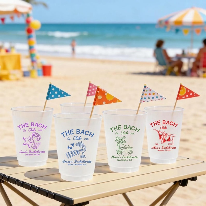 May include: Five clear plastic cups with the text "THE BACH Club" and various graphics, including a seashell, beach umbrella, and palm tree. Each cup has a small flag on a wooden stick. The cups are on a wooden table at the beach.