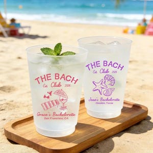 May include: Two frosted plastic cups filled with a clear liquid and ice, garnished with mint. The cups are printed with "THE BACH Est. Club 2026" and bachelorette party details. One cup has a beach-themed design, the other has a starfish and seashell design.