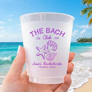 May include: A clear plastic cup with purple text and graphics. The text reads "THE BACH Club Est. 2026" above a starfish, seashell, and conch shell. Below the graphics is "Jean's Bachelorette Houston, Texas".