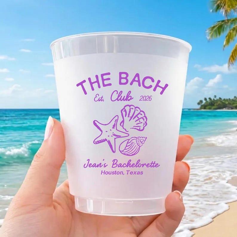 May include: A clear plastic cup with purple text and graphics. The text reads "THE BACH Club Est. 2026" above a starfish, seashell, and conch shell. Below the graphics is "Jean's Bachelorette Houston, Texas".