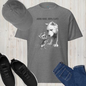 Puede incluir: Una camiseta gris jaspeado con el texto "ZERO FEAR, 100% FLUFF" y un gráfico de un perro pequeño frente a un lobo grande. También se ven una gorra de béisbol gris, zapatillas negras y vaqueros azules.