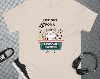 Puede incluir: Camiseta beige con un gráfico de un gato sosteniendo una bebida, rodeado de notas musicales. El texto en la camiseta dice "JUST OUT FOR A PAWSOME EVENING". Zapatillas negras y auriculares también son visibles.