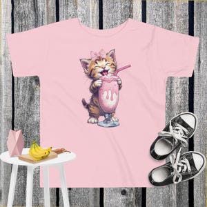 May include: A light pink toddler t-shirt with a cartoon kitten drinking a pink milkshake. The kitten wears a pink bow. Black and white trainers are on the right. A small white table with bananas and a decorative object is on the left.