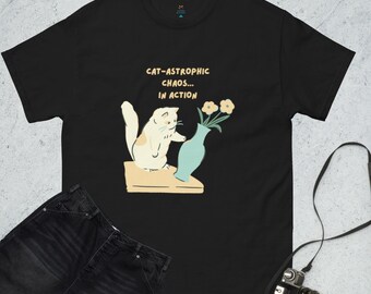 Puede incluir: Camiseta negra con un gato de dibujos animados que alcanza un jarrón azul claro con flores amarillas. El texto "CAT-ASTROPHIC CHAOS... IN ACTION" está encima de la ilustración. Unos vaqueros negros y una cámara también están en la imagen.