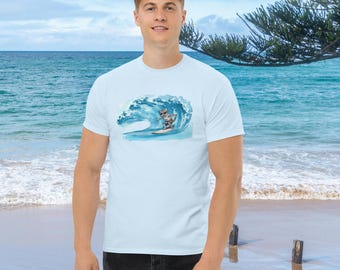 Puede incluir: Camiseta azul claro con una ilustración en acuarela de un gato surfeando una gran ola azul. El gato lleva gafas de sol y practica surf. La camiseta es de color azul claro liso.