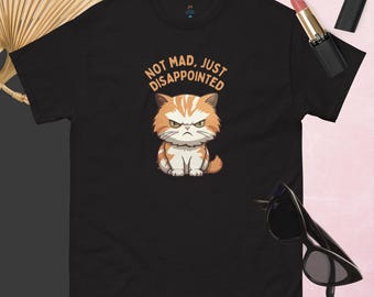 May include: Black t-shirt featuring a cartoon cat with an angry expression. The cat is orange and white, with the text "NOT MAD, JUST DISAPPOINTED" above it. The shirt is displayed with sunglasses and lipstick.