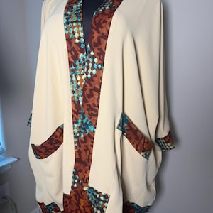 May include: A cream-colored open-front kimono jacket featuring a wide collar, cuffs, and two front pockets accented with a brown and teal geometric pattern. The garment has a loose, flowing silhouette with wide sleeves and a smooth, solid fabric.