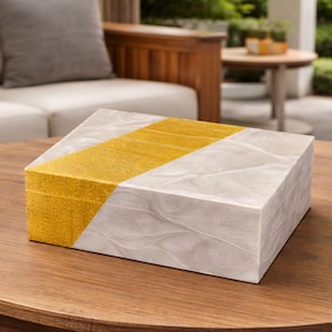 May include: A rectangular decorative box with a diagonal gold stripe on a white background. The box is sitting on a round wooden table. The box has a textured surface and a lid.