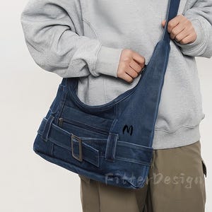 Può includere: Una borsa a tracolla in denim blu con chiusura a patta e dettaglio fibbia. La borsa ha una tasca frontale e una tracolla. La lettera "M" è ricamata sul lato. Il testo "Fitter Design" è visibile.