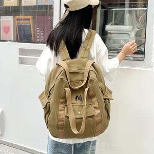 Puede incluir: Una mochila de lona color caqui con múltiples bolsillos y correas. La mochila tiene la letra "M" bordada en la parte delantera. La persona lleva una camisa blanca, vaqueros azul claro y una gorra de béisbol beige.