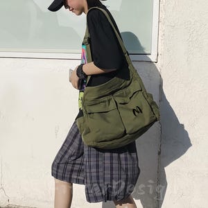 Può includere: Una borsa messenger verde oliva con più tasche frontali e la lettera nera "M" sul lato. La borsa è indossata a tracolla. La persona indossa un berretto nero, una maglietta nera e pantaloncini a quadri.