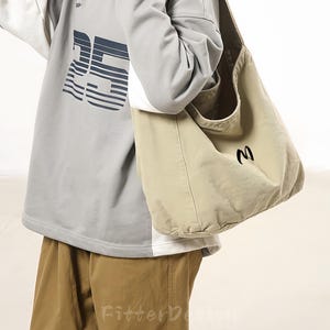 May include: A person wearing a gray sweatshirt with the number "25" printed on the front, paired with brown pants. They are carrying a beige tote bag with a black "M" on it. The text "FitterDesign" is visible on the pants.