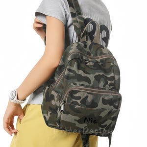 May include: Camouflage-patterned backpack in shades of green, brown, and tan. The backpack has a front pocket with a zipper and adjustable straps. The word "Mia" is printed on the front. The backpack is designed for everyday use.