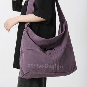 Può includere: Una grande borsa a tracolla viola polveroso con una lunga tracolla. La borsa ha il nome "Emma" ricamato, insieme al testo "FitterDesign". La borsa è realizzata in un tessuto robusto e si indossa a spalla.