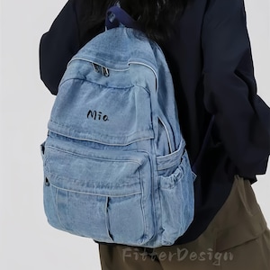 May include: Light blue denim backpack with multiple pockets and the name "Mia" in black script. The backpack has a top handle and adjustable straps. The design includes a white zipper and a white trim. The backpack is worn over the shoulder.