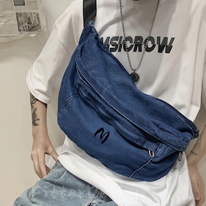 Puede incluir: Bolso cruzado de mezclilla azul con una correa negra. El bolso tiene forma curva y presenta la letra "M" en negro en la parte delantera. La persona lleva una camiseta blanca con la palabra "MUSICROW" impresa.