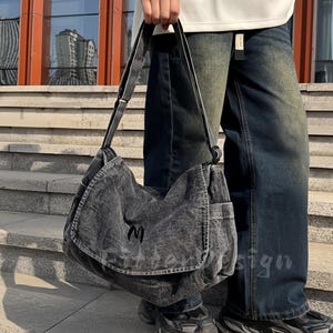 Può includere: Una borsa messenger in denim grigio con patta e tracolla. La borsa presenta una piccola "M" ricamata sul davanti. La persona nell'immagine indossa una felpa bianca e jeans blu.