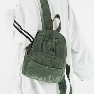 Może przedstawiać: Zielony sztruksowy plecak z przednią kieszenią i regulowanymi paskami. Plecak ma małe haftowane "M" z przodu i górny uchwyt. Torba jest noszona na ramieniu.