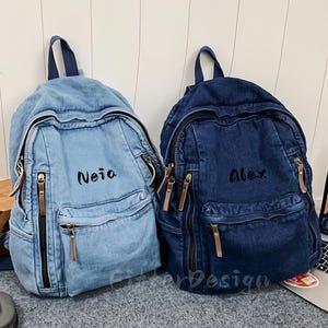 Könnte beinhalten: Zwei Jeans-Rucksäcke, einer hellblau und einer dunkelblau, mit goldenen Reißverschlüssen und den Namen "Neia" und "Alex" darauf gestickt. Die Rucksäcke haben ein klassisches Design mit einem oberen Griff und einer Vordertasche.