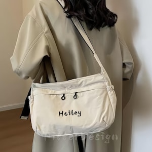 Könnte beinhalten: Eine beige Umhängetasche mit einem schwarzen Schulterriemen. Die Tasche hat zwei schwarze Knopfverschlüsse und den Namen "Hailey" in schwarzer Schrift. Die Tasche ist aus einem weichen, hellfarbigen Material gefertigt.