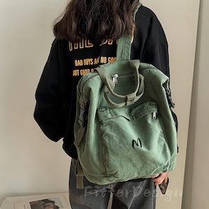 Może przedstawiać: Szałwiowy zielony płócienny plecak z wieloma kieszeniami i górnym uchwytem. Plecak ma czarne haftowane "M" na przedniej kieszeni i regulowane paski. Osoba ma na sobie czarną bluzę z kapturem ze złotym tekstem na plecach.