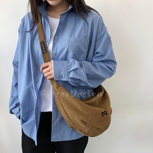 May include: A light blue button-down shirt with a white undershirt and a brown shoulder bag. The bag has a curved shape and a black "M" on the front. The bag's strap is brown.