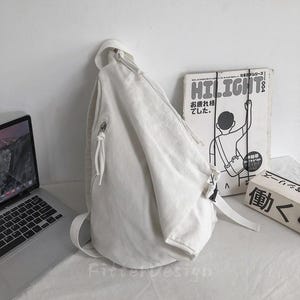 Puede incluir: Una mochila bandolera de lona blanca con una sola correa para el hombro y un bolsillo con cremallera. La bolsa est&aacute; apoyada contra una pared blanca, con un port&aacute;til y una revista vintage en el fondo.