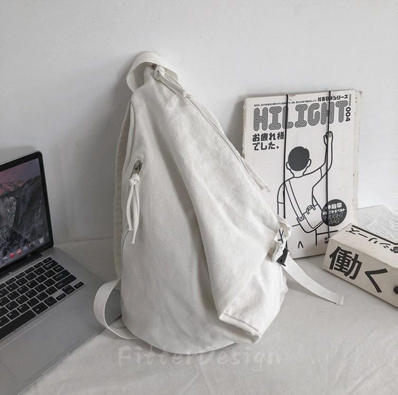 Puede incluir: Una mochila bandolera de lona blanca con una sola correa para el hombro y un bolsillo con cremallera. La bolsa est&aacute; apoyada contra una pared blanca, con un port&aacute;til y una revista vintage en el fondo.