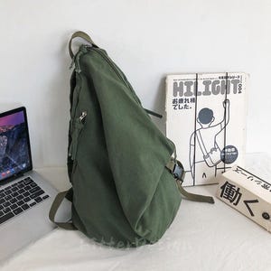 Puede incluir: Mochila bandolera de lona verde oliva con un bolsillo con cremallera y una correa ajustable. La bolsa est&aacute; al lado de un port&aacute;til y un libro con la palabra "HILIGHT" en la portada.
