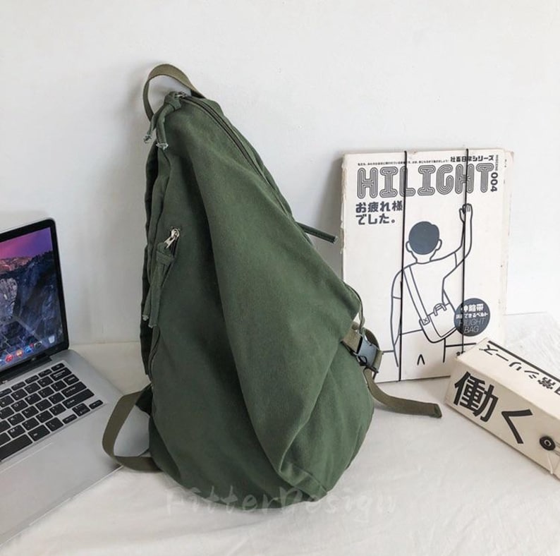 Puede incluir: Mochila bandolera de lona verde oliva con un bolsillo con cremallera y una correa ajustable. La bolsa est&aacute; al lado de un port&aacute;til y un libro con la palabra "HILIGHT" en la portada.