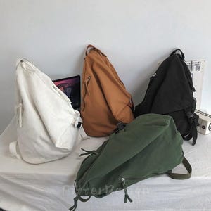 Puede incluir: Cuatro mochilas bandoleras de lona en blanco, marr&oacute;n, negro y verde oliva. Cada bolsa tiene una sola correa para el hombro, un compartimento principal con cremallera y correas ajustables. Las bolsas se muestran sobre un fondo blanco.