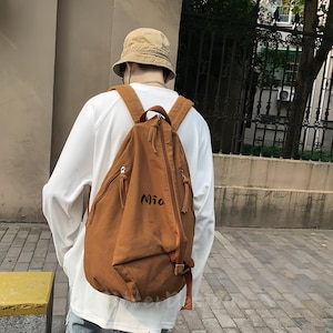 Puede incluir: Una mochila marr&oacute;n con el nombre "Mia" impreso en la parte delantera. La mochila tiene dos bolsillos con cremallera y correas ajustables. La persona de la imagen lleva una camisa blanca de manga larga y un sombrero de pescador beige.