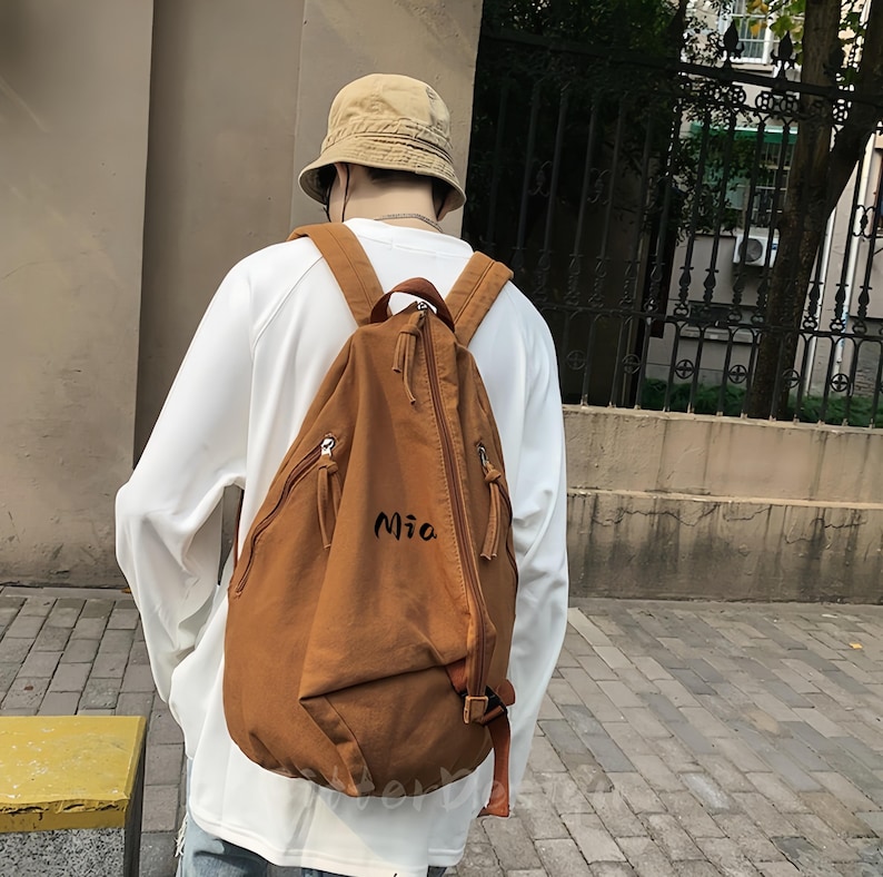 Puede incluir: Una mochila marr&oacute;n con el nombre "Mia" impreso en la parte delantera. La mochila tiene dos bolsillos con cremallera y correas ajustables. La persona de la imagen lleva una camisa blanca de manga larga y un sombrero de pescador beige.