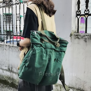 Op de afbeelding: Een groene canvas rugzak met een ritssluiting en een flap. De rugzak heeft een schouderriem en een handvat aan de bovenkant. De letter "M" is op de voorkant geborduurd. De tekst "Fitterdesign" staat onderaan.