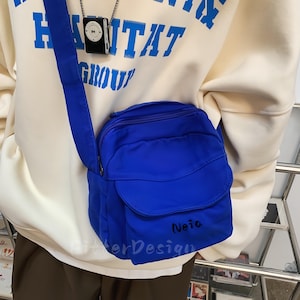 以下が含まれることがあります： 鮮やかな青色のクロスボディバッグ。フラップポケットとショルダーストラップが付いています。バッグは丸みを帯びた形で、手書きの「Neia」と「AlterDesign」の文字が特徴です。クリーム色のスウェットシャツの上から着用されています。
