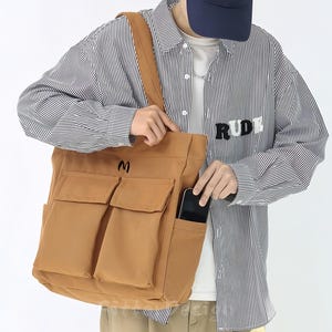 Peut inclure: Un sac fourre-tout en toile beige avec deux poches avant et une poche latérale. Le sac a une bandoulière et un "M" noir brodé sur le devant. Un smartphone est visible dans la poche latérale.
