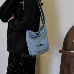 Puede incluir: Bolso de hombro de mezclilla azul claro con la palabra "Dolly" bordada en negro. El bolso tiene un bolsillo frontal y una correa larga. El bolso se lleva al hombro.