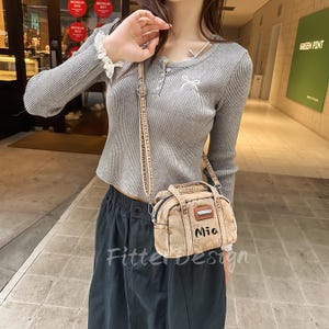 May include: A grey, long-sleeved ribbed top with a small bow detail and a small tan handbag with the name "Mia" on it. The handbag has a long strap. The text "Fitterdesign" is visible on the bottom of the image.